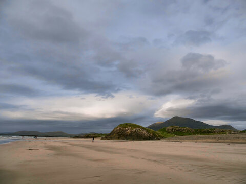 Bare sandy beach stretches toward grassy, rounded hills under cloudy sky. Hills rise gently with patches of green and exposed rock. Scene feels quiet and serene, perfect for nature or travel themes