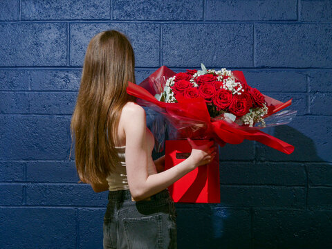 Woman holds vibrant red roses bouquet against blue brick wall. She stands with back turned, suggesting anticipation or surprise. Perfect for Valentine's Day, anniversaries, or romantic gestures