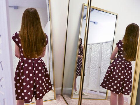 Young girl with long hair stands before tall, golden-framed mirrors. She wears a polka-dot dress, examining her reflection. Her posture suggests quiet curiosity and self-discovery