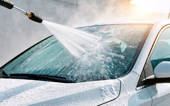 Worker uses high pressure washer to clean car windshield. Strong water spray hits glass surface of silver automobile.
