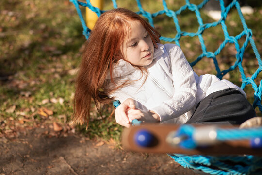 Red-haired young girl relaxing on a net swing in autumn park
