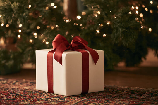 White Gift with red ribbon on patterned rug before glowing Christmas tree creating classic cozy holiday scene