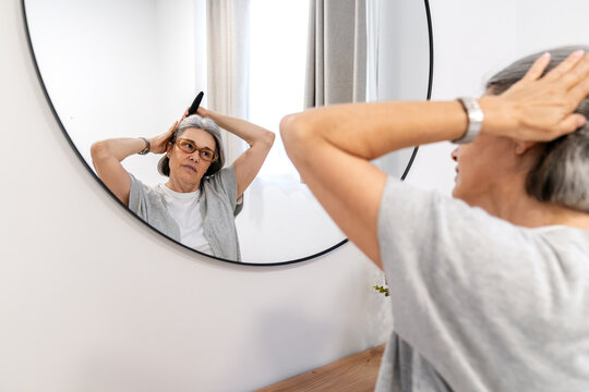 Senior woman in glasses brushing her gray hair while checking her reflection in a round mirror at home