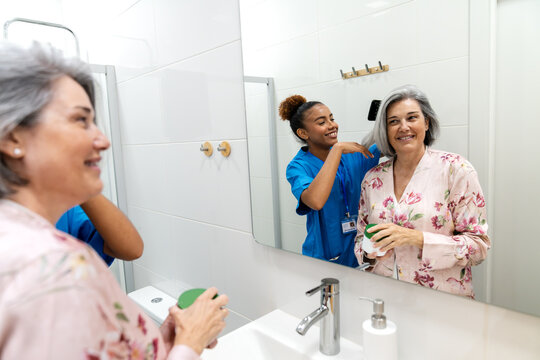 Home health nurse assisting an older woman with grooming, gently arranging her hair as they share a warm moment at the bathroom mirror