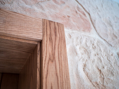 Close-up of a solid oak wooden door frame with natural wood grain and detailed corner joinery beside a decorative gypsum plaster wall imitating stone. Rustic interior finishing detail.