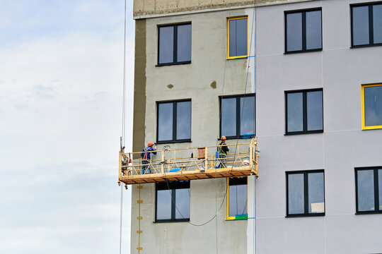 Construction workers on suspended cradle platform plastering exterior wall facade of multi story residential building. Builders on suspended scaffolding cradle finishing and plastering facade wall