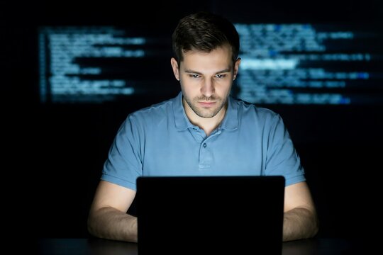 Focused young man coding on laptop with blue digital programming code displayed on dark background screen monitor