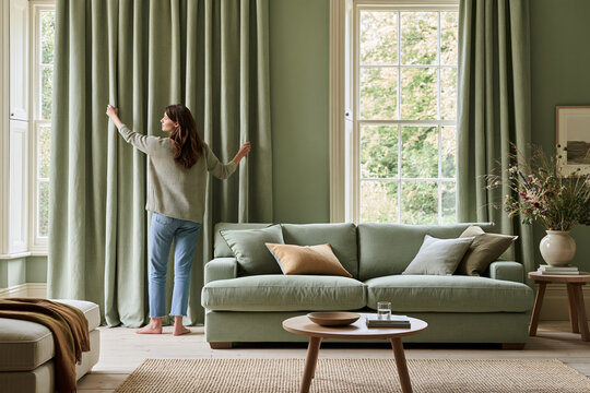 Woman opening sage green curtains in moss color drenching living room, comfortable sofa with tonal decor, morning light