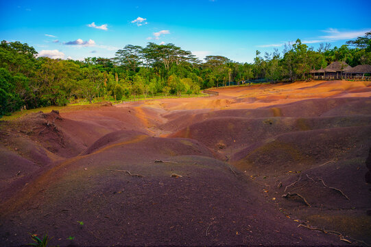 Multicolored sand dunes at Chamarel Seven Colored Earth Geopark, with a wooden lodge set among lush tropical forest under a bright blue sky in Mauritius.