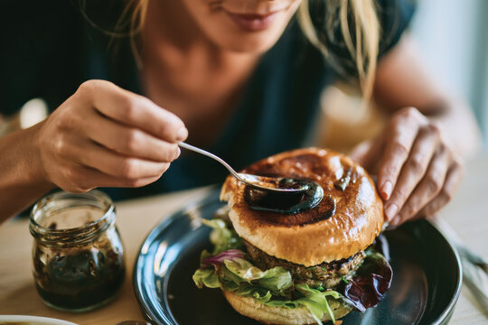 A woman enhances her gourmet burger with a luscious sauce, highlighting the art of creating an exquisite culinary experience with each bite.