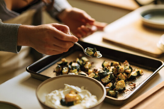 A person elegantly garnishes roasted vegetables on a baking tray with a dollop of yogurt, showcasing a blend of textures and flavors in a kitchen setting.