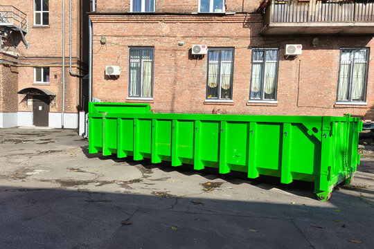Bright green industrial waste container standing in courtyard beside a brick building on a sunny day, representing urban waste management and environmental responsibility.