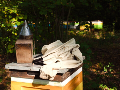 Beekeeper protective jacket with veil hood and honey bees crawling on fabric, laid on wooden beehive beside old metal bee smoker, apiary in shady summer forest, beekeeping workwear, apiculture.