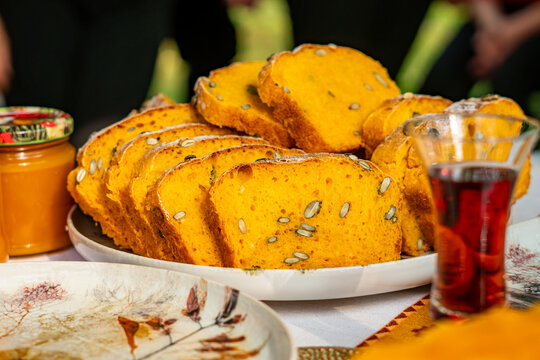 Close up shot of thick slices of homemade yellow pumpkin bread with pumpkin seeds arranged on a plate, displayed on an autumn table setting next to a glass of tea and a jar of preserves in natural lig